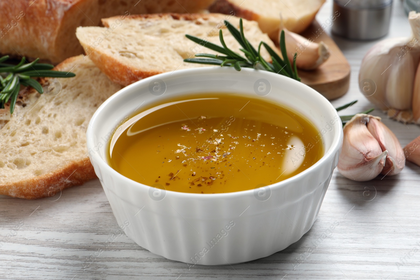 Photo of Bowl of fresh oil, bread, rosemary and garlic on white wooden table, closeup