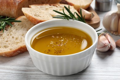 Photo of Bowl of fresh oil, bread, rosemary and garlic on white wooden table, closeup