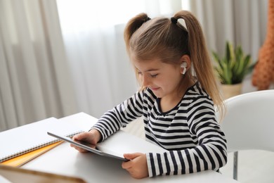 Photo of Adorable little girl doing homework with tablet and earphones at table indoors