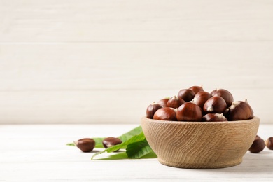 Photo of Fresh sweet edible chestnuts in bowl on white wooden table. Space for text