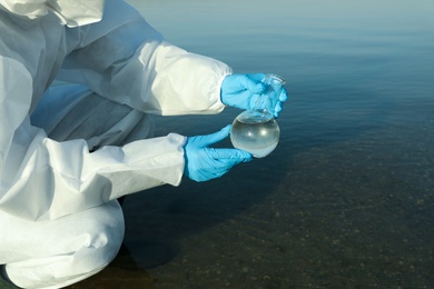 Scientist in chemical protective suit with florence flask taking sample from river for analysis, closeup