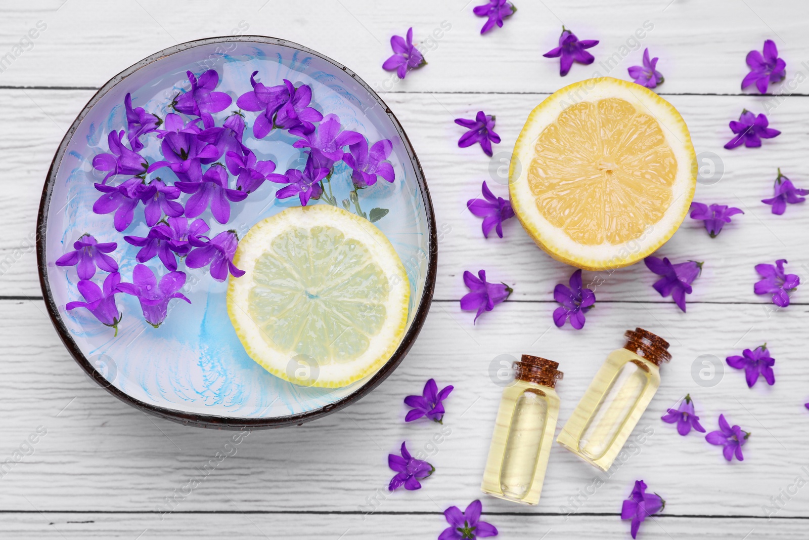 Photo of Bowl with water, lemon, purple flowers and bottles of essential oils on white wooden table, flat lay. Spa composition