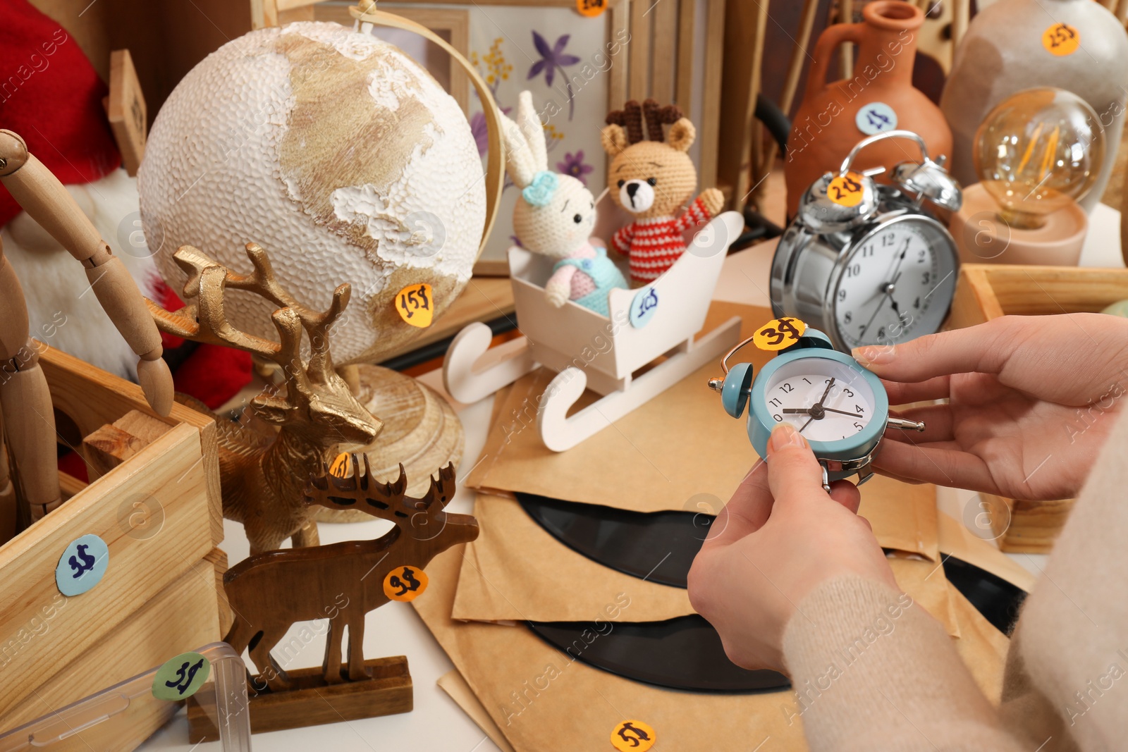 Photo of Woman holding alarm clock near table with many different stuff indoors, closeup. Garage sale