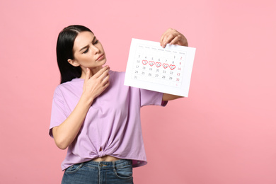 Pensive young woman holding calendar with marked menstrual cycle days on pink background