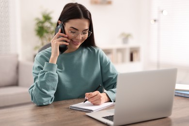 Photo of Young woman talking on smartphone during webinar at table in room