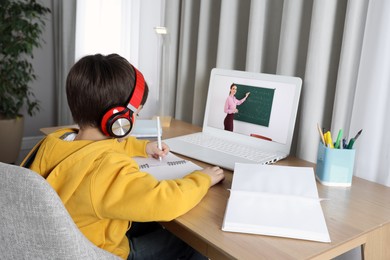 Image of E-learning. Little boy taking notes during online lesson at wooden table indoors