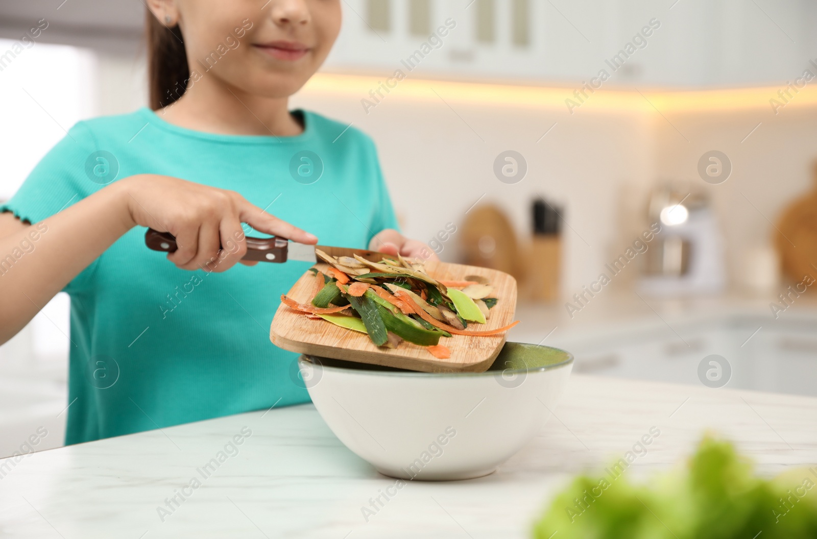 Photo of Little girl with cutting board and knife scraping  vegetable peels into bowl on kitchen table, closeup