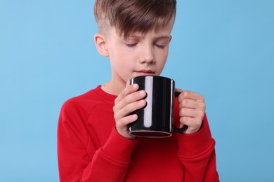 Photo of Cute boy drinking beverage from black ceramic mug on light blue background
