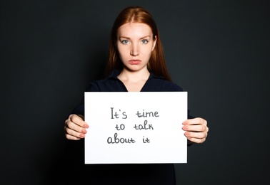 Young woman holding card with words IT'S TIME TO TALK ABOUT IT against dark background
