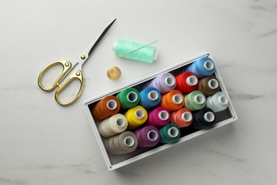 Photo of Box with colorful sewing threads, scissors and thimble on white marble table, flat lay