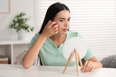 Suffering from allergy. Young woman looking in mirror and scratching her face at white table indoors
