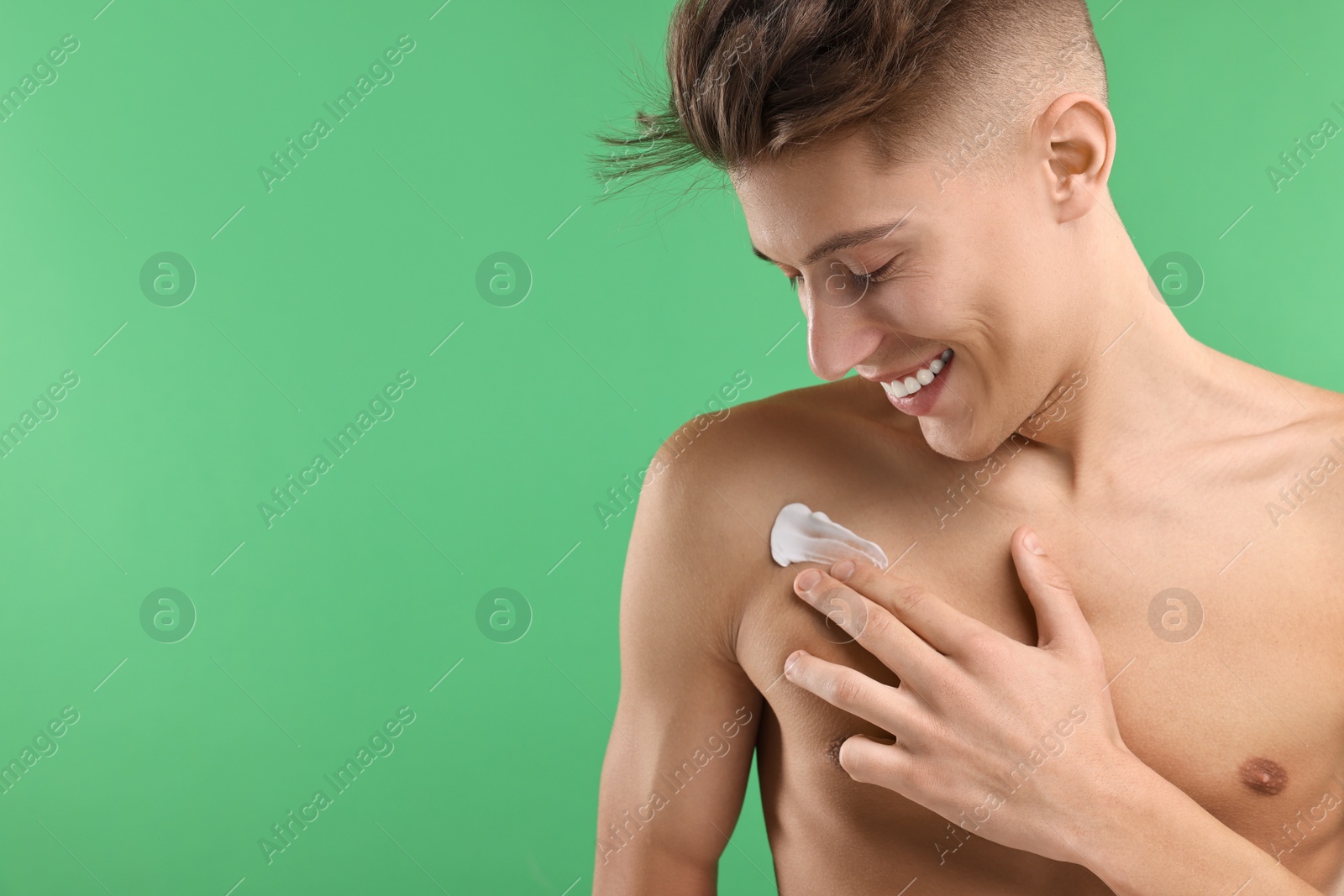 Photo of Handsome man applying moisturizing cream onto his shoulder on green background. Space for text