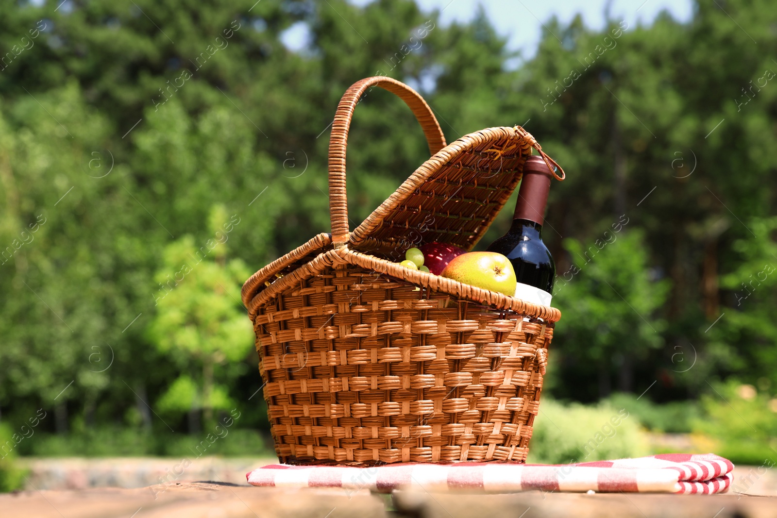 Photo of Picnic basket with fruits, bottle of wine and checkered blanket on wooden table in garden