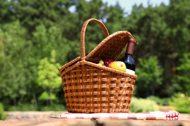Picnic basket with fruits, bottle of wine and checkered blanket on wooden table in garden