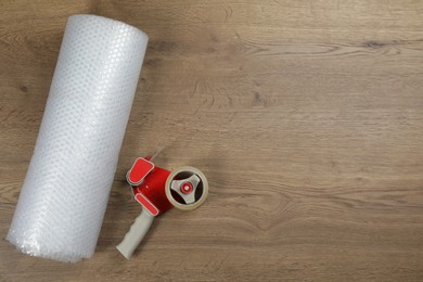 Photo of Bubble wrap roll and tape dispenser on wooden background, flat lay. Space for text