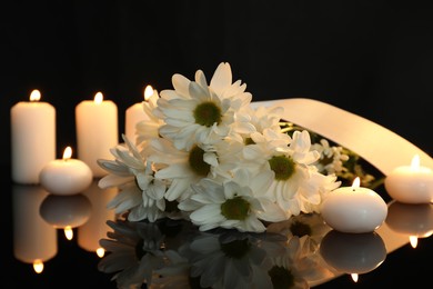 Photo of White chrysanthemum flowers and burning candles on black mirror surface in darkness. Funeral symbols