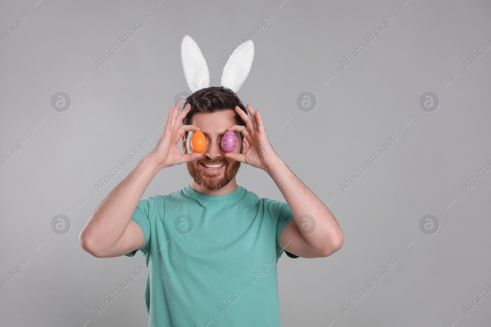 Photo of Happy man in cute bunny ears headband covering eyes with Easter eggs on light grey background