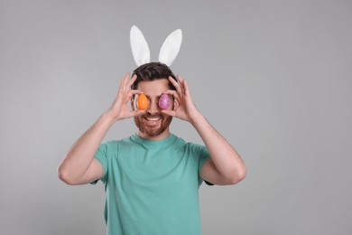 Photo of Happy man in cute bunny ears headband covering eyes with Easter eggs on light grey background