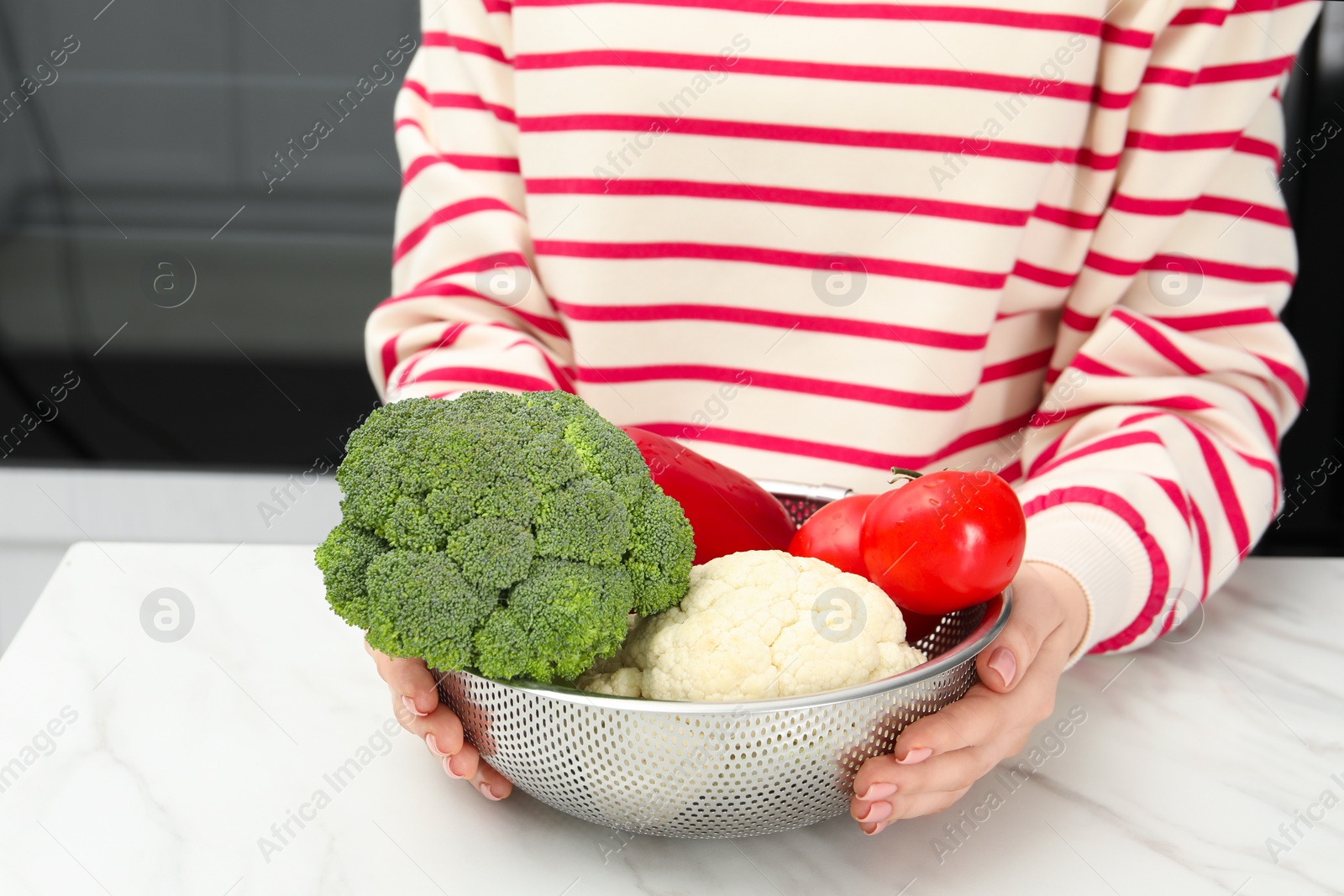 Photo of Woman holding colander with fresh vegetables at white marble table in kitchen, closeup