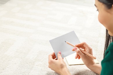 Photo of Woman writing message in greeting card on carpet in room, closeup. Space for text