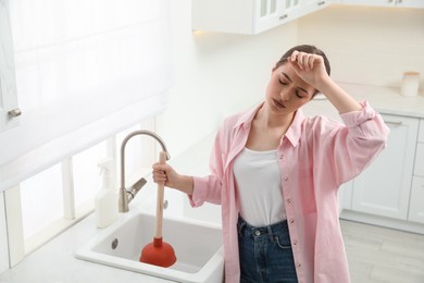 Photo of Tired young woman using plunger to unclog sink drain in kitchen