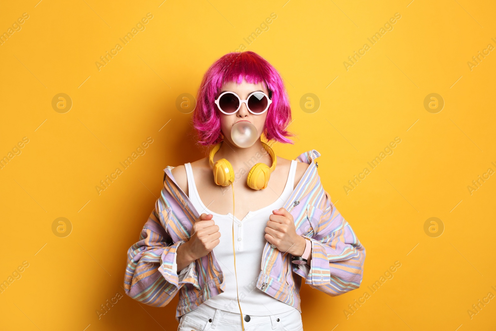 Photo of Fashionable young woman in colorful wig with headphones blowing bubblegum on yellow background