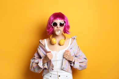 Photo of Fashionable young woman in colorful wig with headphones blowing bubblegum on yellow background
