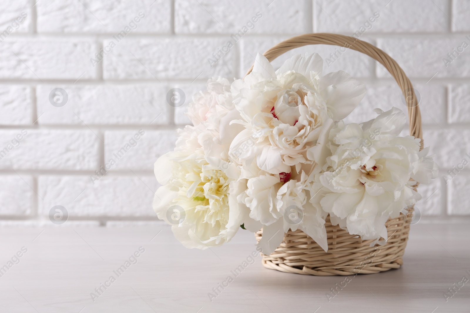 Photo of Beautiful peonies in wicker basket on white table near brick wall