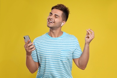 Happy young man with smartphone listening to music through wireless earphones on yellow background