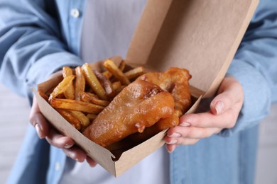 Photo of Woman holding fish and chips in paper box near white brick wall, closeup