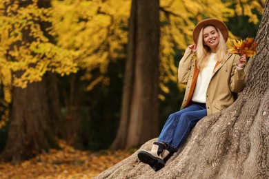 Photo of Happy woman with dry autumn leaves sitting on tree trunk outdoors. Space for text