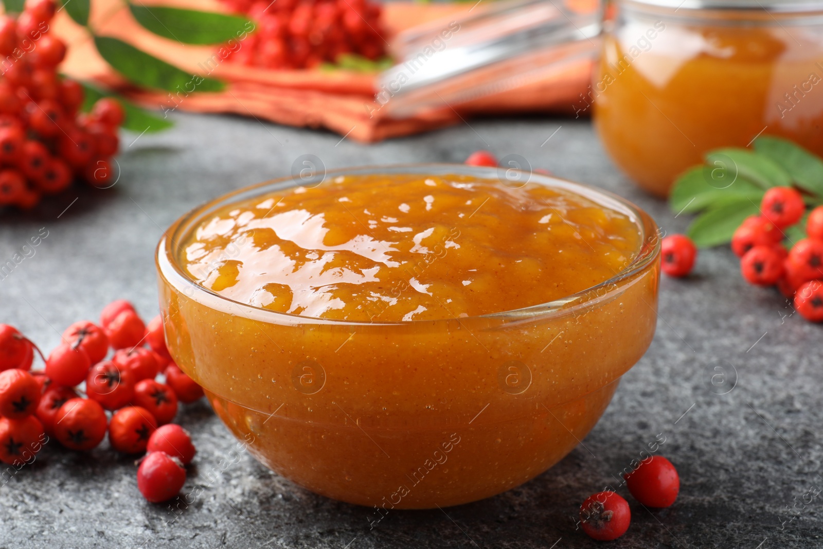 Photo of Delicious rowan jam in glass bowl and berries on grey table, closeup