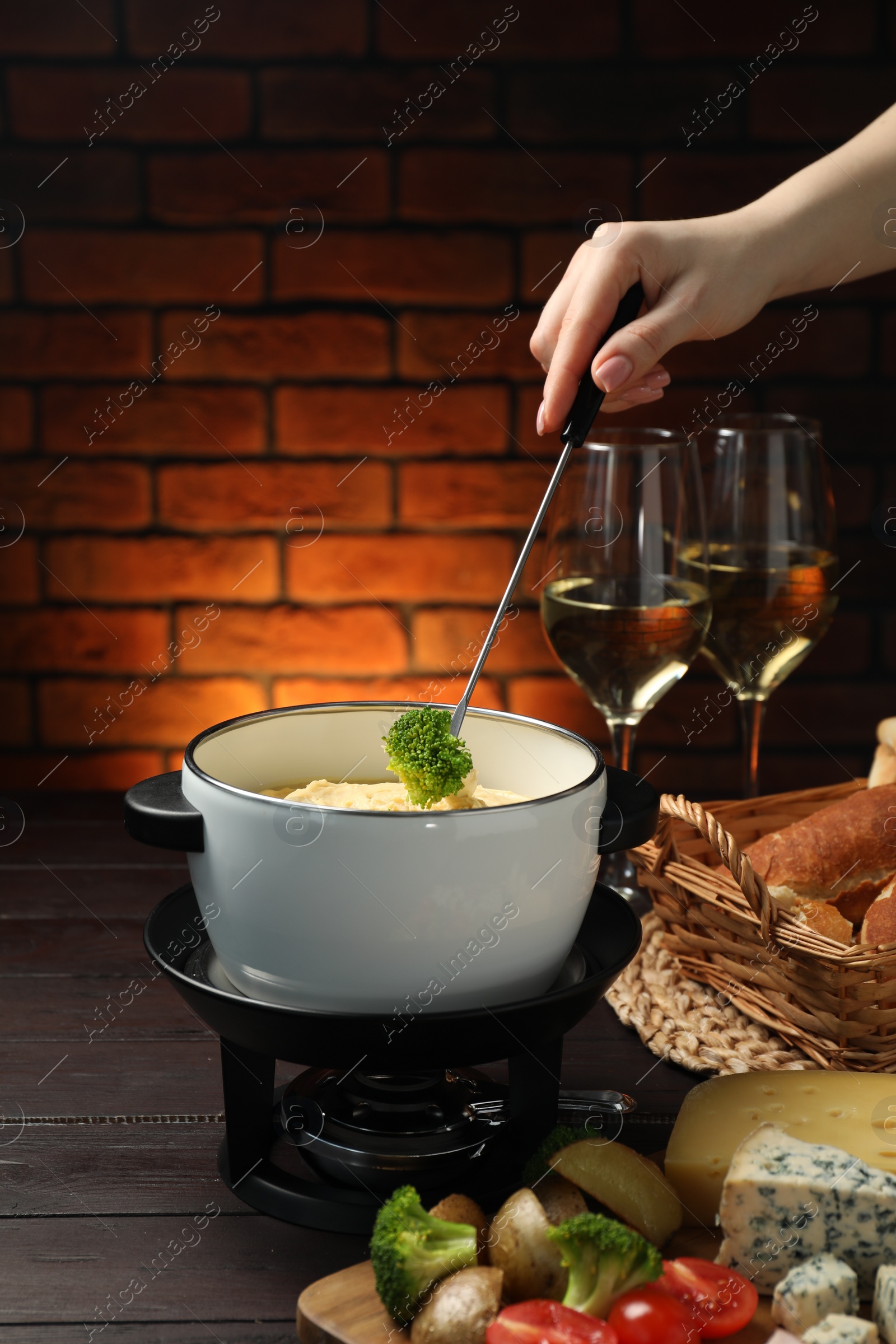 Photo of Woman dipping piece of broccoli into fondue pot with melted cheese at wooden table with wine and snacks, closeup