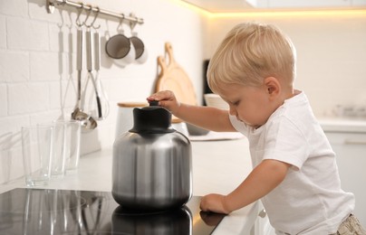 Photo of Curious little boy playing with kettle on electric stove in kitchen