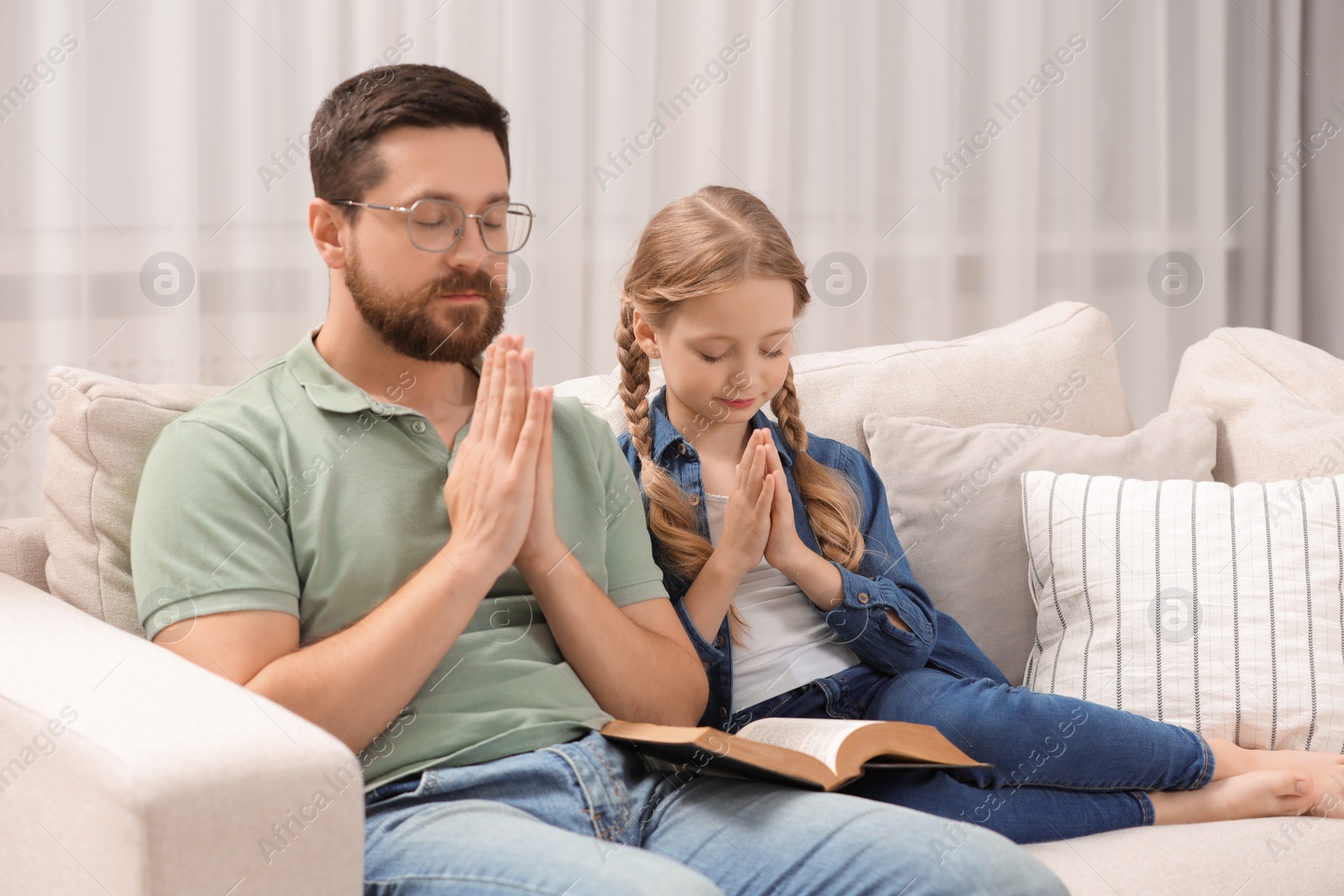 Photo of Girl and her godparent praying over Bible together on sofa at home