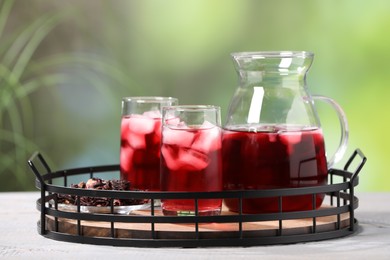 Photo of Refreshing hibiscus tea with ice cubes and dry roselle flowers on white wooden table against blurred green background