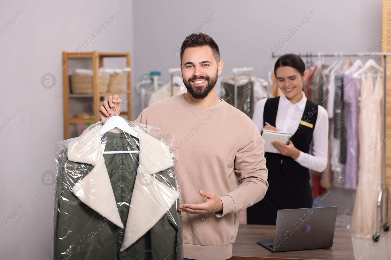 Photo of Dry-cleaning service. Happy man holding hanger with jacket indoors. Worker taking notes at workplace