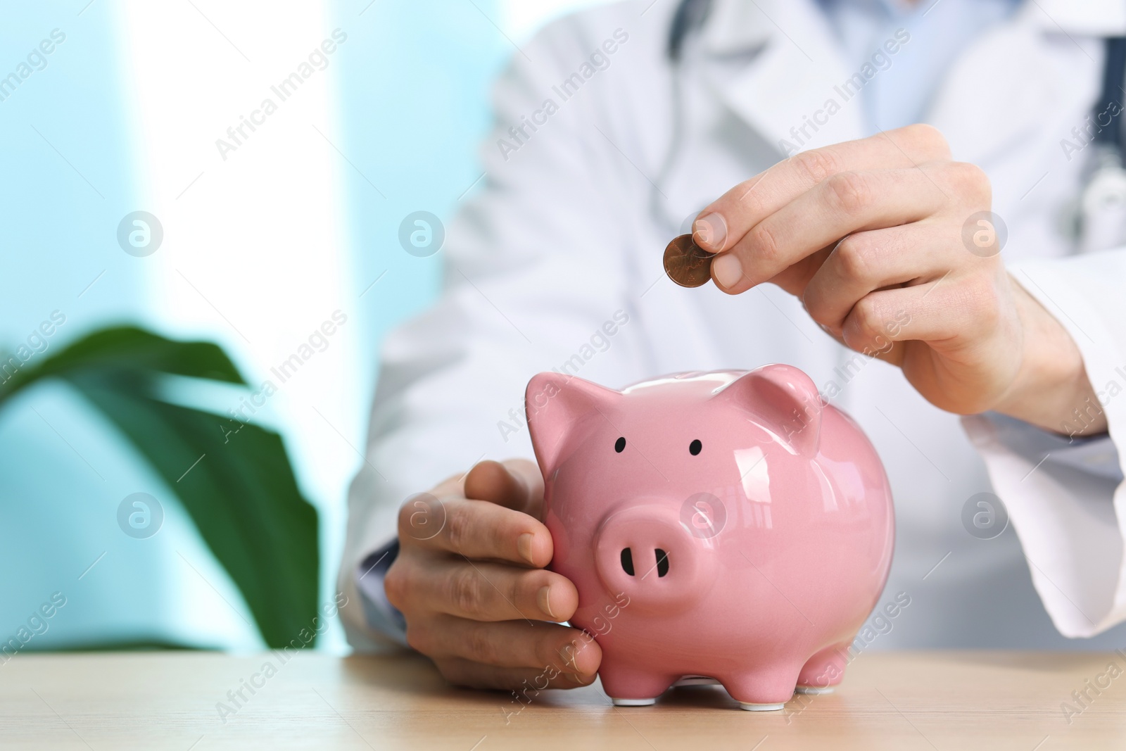 Photo of Doctor putting coin into piggy bank at wooden table, closeup