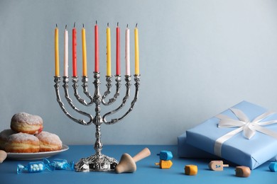 Hanukkah celebration. Menorah with burning candles, dreidels, donuts and gift boxes on light blue table, space for text