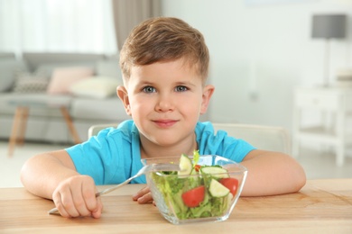 Adorable little boy eating vegetable salad at table in room
