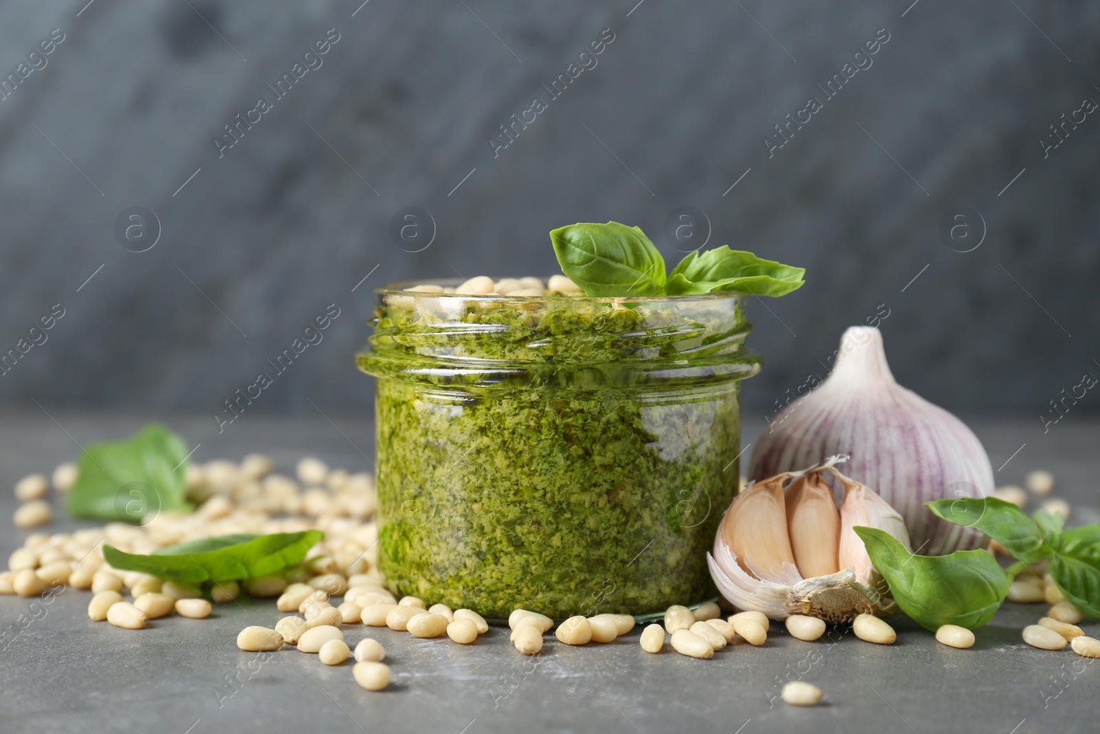 Photo of Jar of delicious pesto sauce and ingredients on grey table, closeup