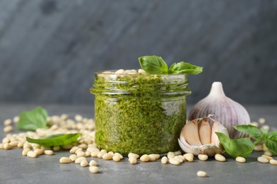 Photo of Jar of delicious pesto sauce and ingredients on grey table, closeup