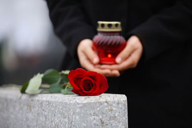 Woman holding candle near grey granite tombstone outdoors, closeup. Funeral ceremony