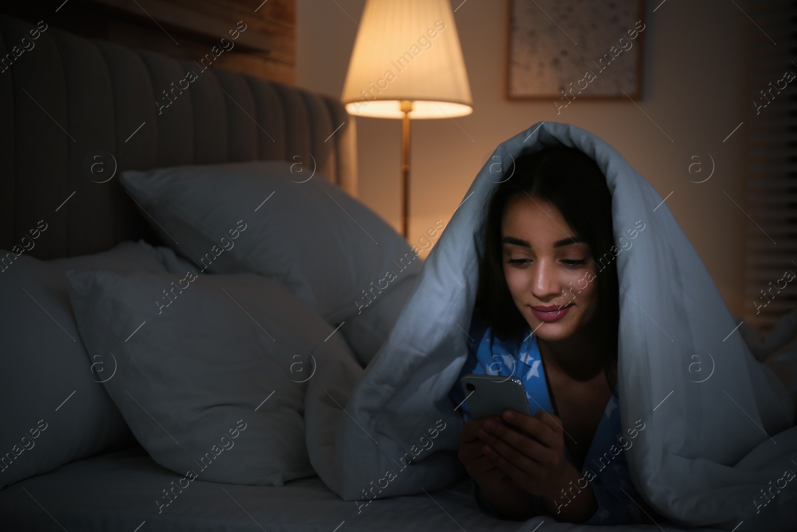 Photo of Happy woman using mobile phone under blanket in dark bedroom