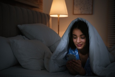 Photo of Happy woman using mobile phone under blanket in dark bedroom