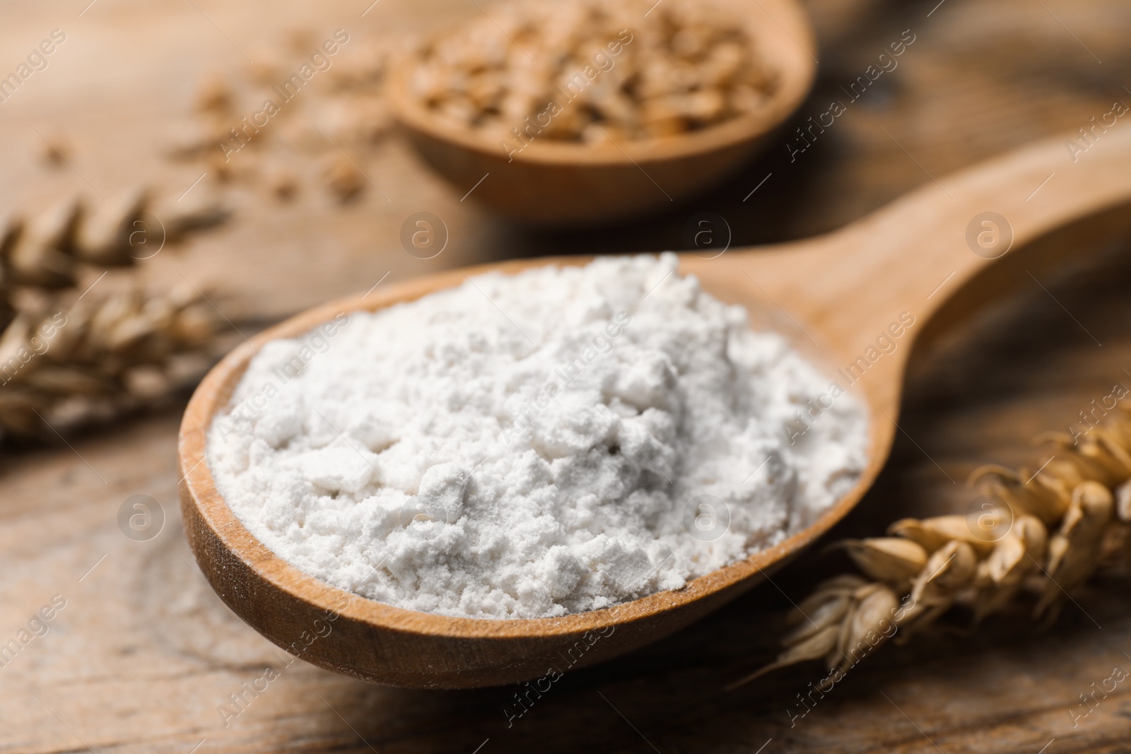 Photo of Spoon with wheat flour and spikes on wooden table, closeup