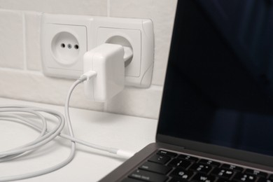 Photo of Charging laptop with power adapter in electrical socket on white table, closeup