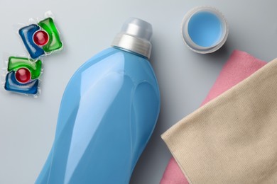 Photo of Bottle of fabric softener, laundry capsules and stacked clean clothes on light background, flat lay