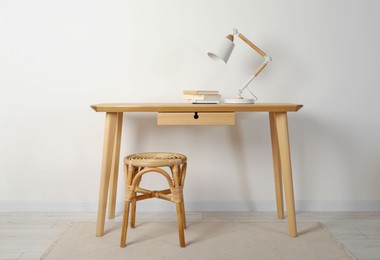 Photo of Wooden table with stylish desk lamp and wicker stool near white wall indoors