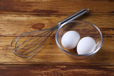 Photo of Metal whisk and eggs in bowl on wooden table, closeup